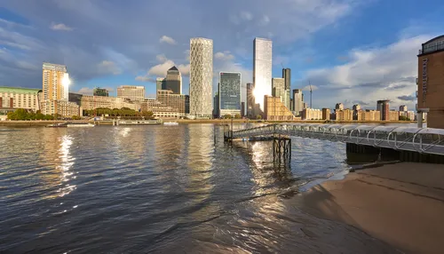 This urban landscape photograph captures Canary Wharf Doubletree in London, United Kingdom during a sunny autumn afternoon. The image features the River Thames in the foreground, with its rippling water reflecting the sunlight and the modern architecture of the Canary Wharf skyline, including prominent office towers. The sun casts bright reflections on the glass facades of the skyscrapers, highlighting the dynamic atmosphere of this urban district. The view showcases the blending of riverside elements and high-rise structures typical of Canary Wharf, making the photograph an exemplary representation of London's iconic financial centre along the Thames.